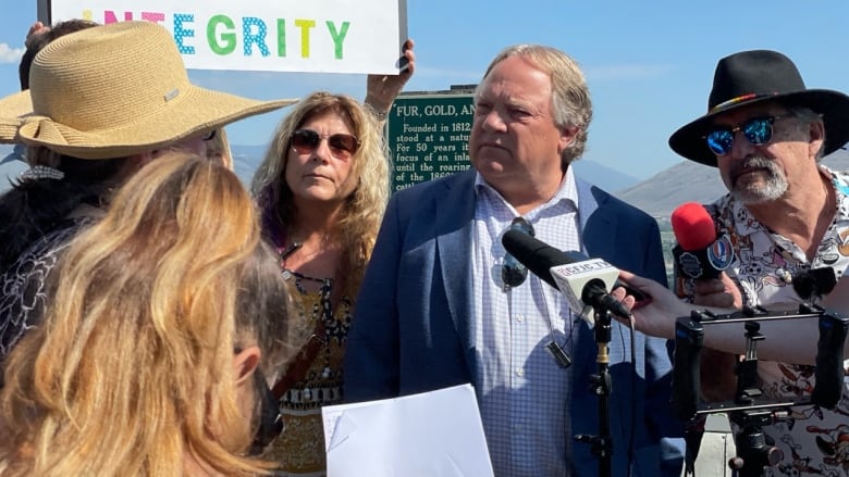 A group of reporters speak to a man and hold mics in front of him, while a woman looks on and a person holds up a sign reading 'Mayor has integrity' in the background.