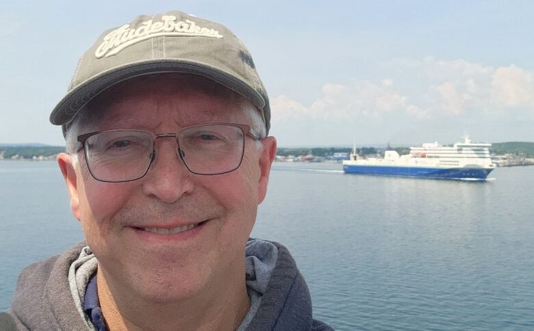 A smiling man takes a selfie in front of the ocean. A ferry vessel travels in the background.