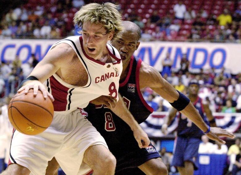 A man in a white Team Canada jersey dribbles a basketball around an American defender. 