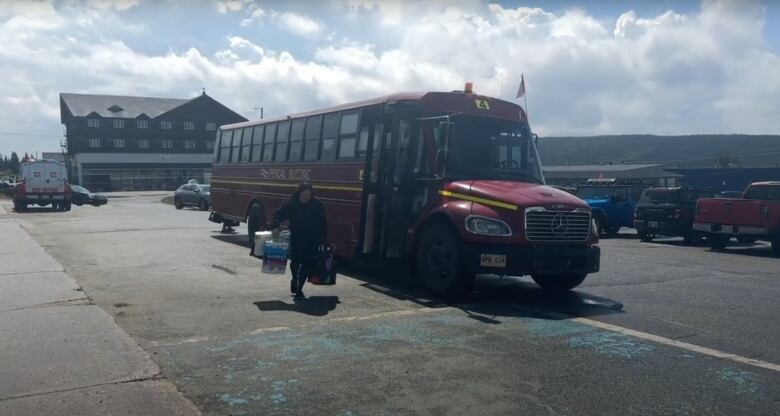 A woman carrying a bag of groceries and a bundle of toilet paper walks out of a red bus.