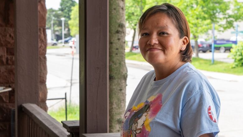 A person is seen standing on a porch, smiling. The road and some green trees can be seen in the background.