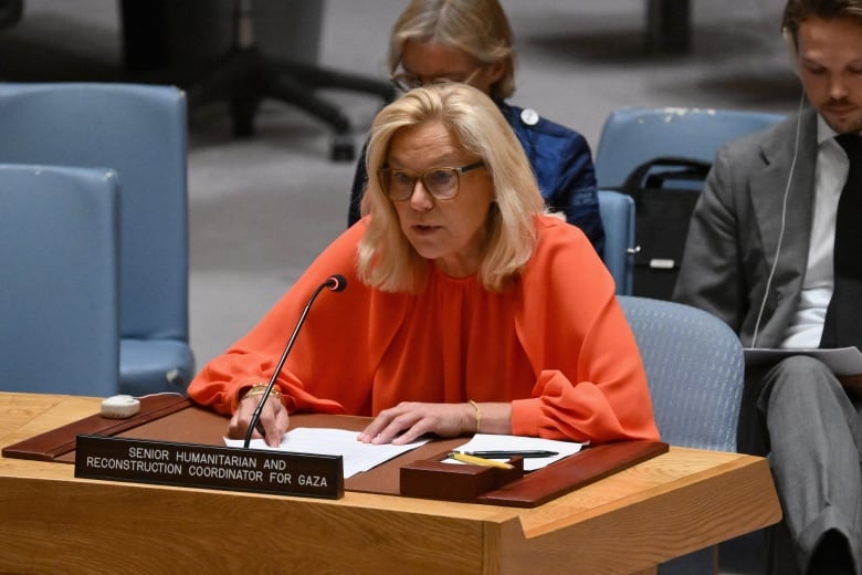 A woman in orange sits at a wooden table with a microphone. In front of her is a black placard that says 'SENIOR HUMIANITARIAN AND RECONSTRUCTION COORDINATOR FOR GAZA.'