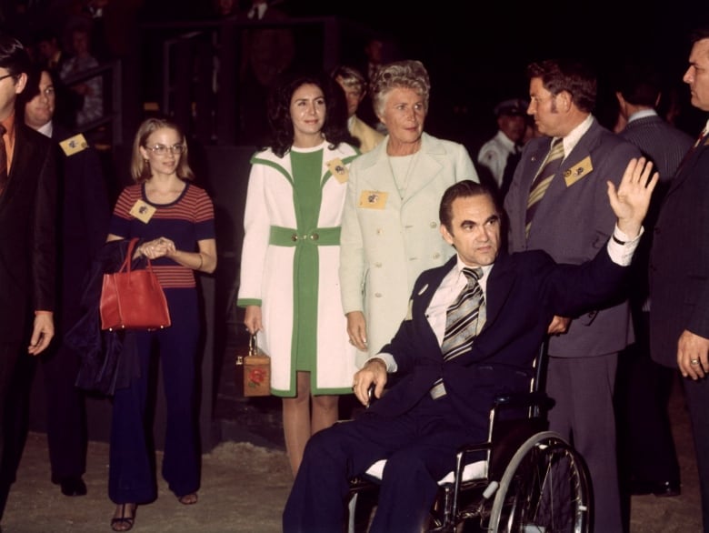 A man in a suit and tie sitting in a wheelchair waves to a person off camera at an event. Near him are men and women standing