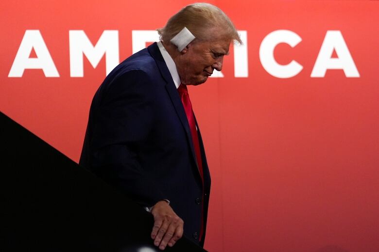 A person with a bandaged ear walk down a flight of stairs as the word 'America' is seen on the wall behind them.