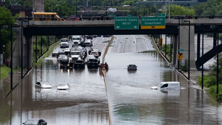 Several cars are seen partially submerged in water on a roadway, with traffic stopped ahead of the water.