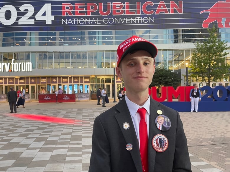 A young man wearing a red Make America Great Again hat poses outside an arena with signage for the Republican National Convention.