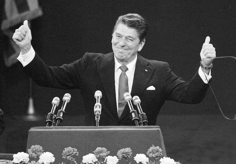 A man in a suit, standing at a lectern, gives a thumbs-up gesture with each hand. 