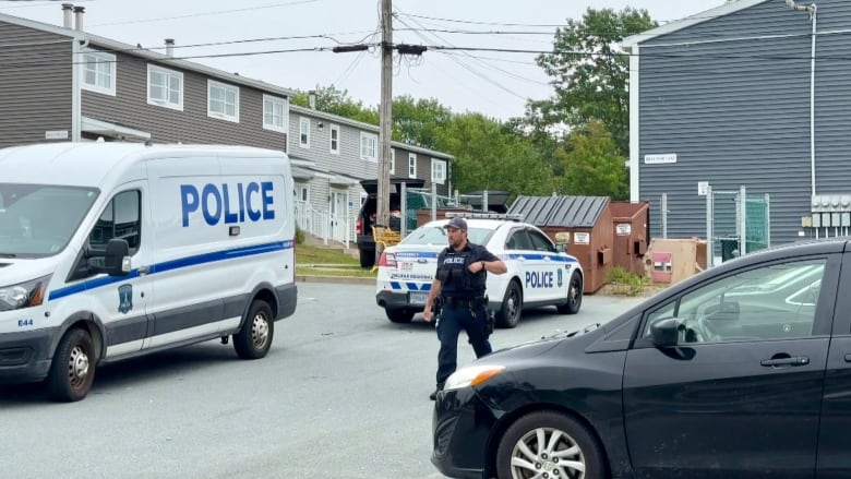 A cop walks down a street. Two police vehicles are visible to his left side and a row of townhouses can be seen in the background.