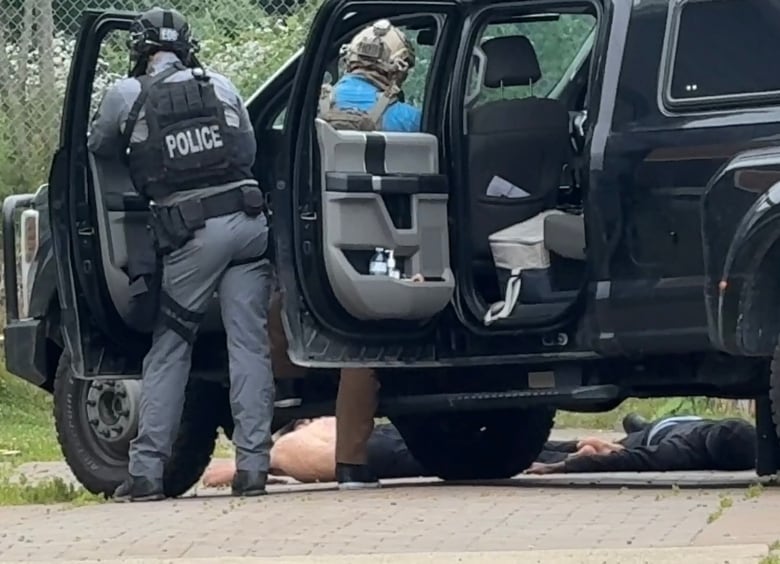 A black vehicle is shown with the doors open and police officers standing in front. Two people can be seen in the background lying on the ground.