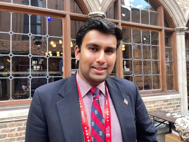 A young man wearing a suit, checkered shirt and a lanyard around his neck smiles.