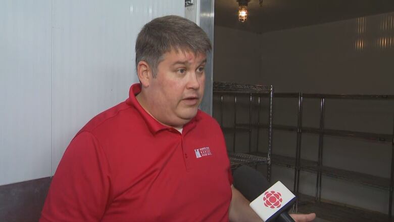 A man in a red shirt stands beside an empty walk-in fridge and talks into a reporter's microphone.