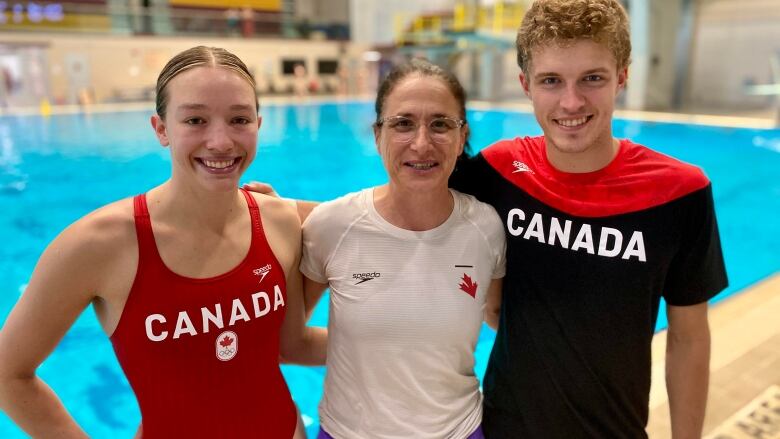 A woman in a red one piece swimsuit, an older woman in a white T-shirt, and a blond, curly-haired man in a Canada shirt link arms while standing in front of a pool. 