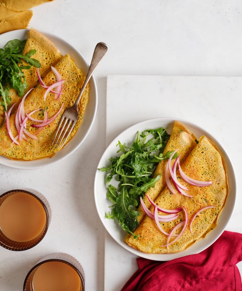 Two plates of thin, folded pancakes with a side of greens and topped with pickled sliced onions.