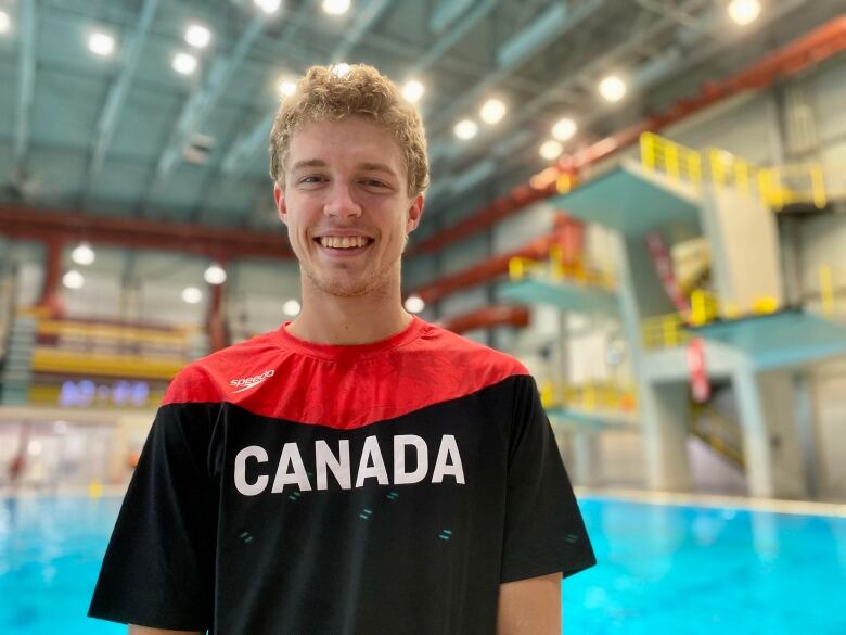 A smiling young man in a black and red Canada T-shirt stands in front of diving boards at a pool.