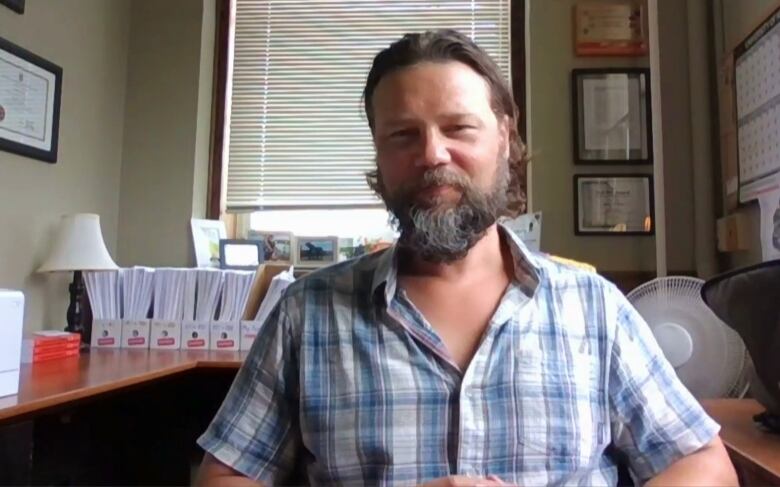 Man with beard in blue plaid shirt sits at desk. 