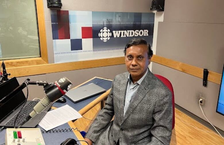 A man in a grey suit sits behind a desk in a radio studio. There is a microphone in front of him and the word 