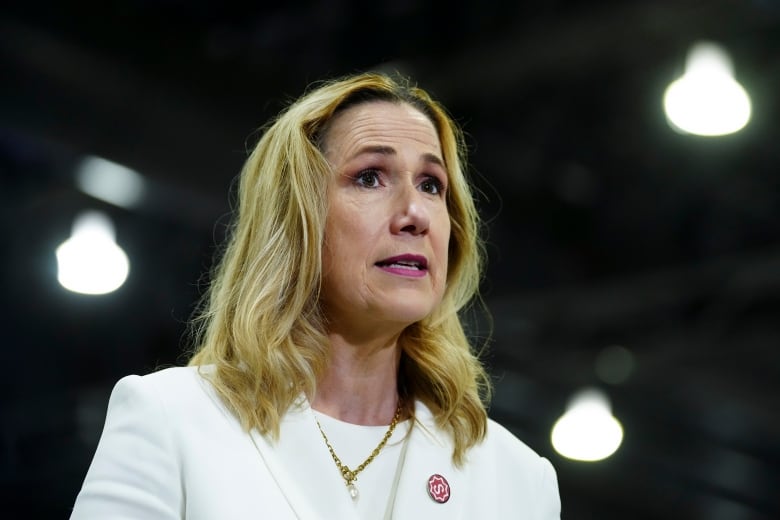 A woman wearing a white jacket is shown speaking during an event.