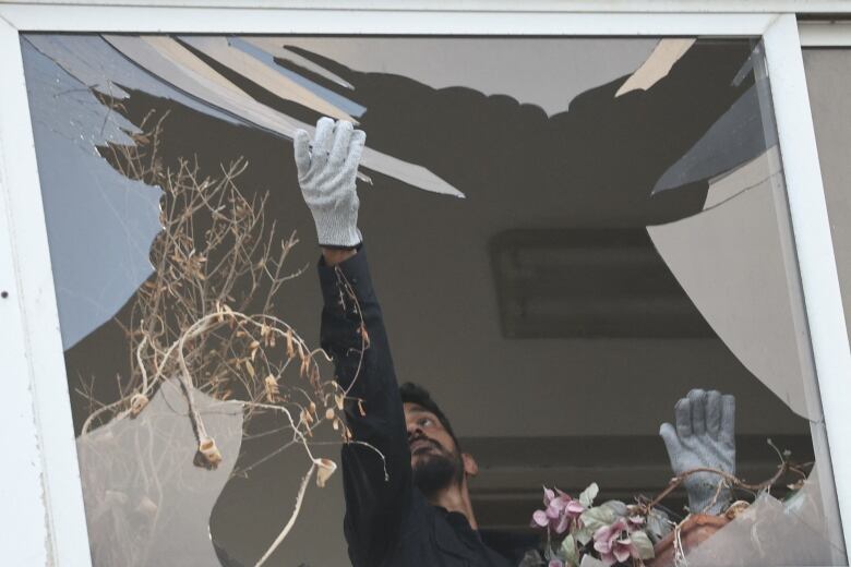 A bearded man wearing gloves reaches up with his arm to grab a shard of glass in a broken window frame.