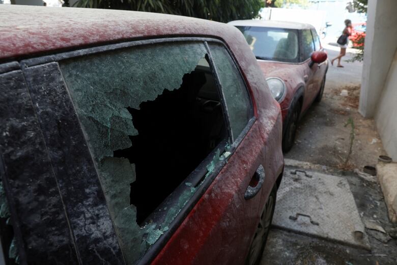 A closeup of a red vehicle is shown with is window blown out and dust and debris on and around the car.