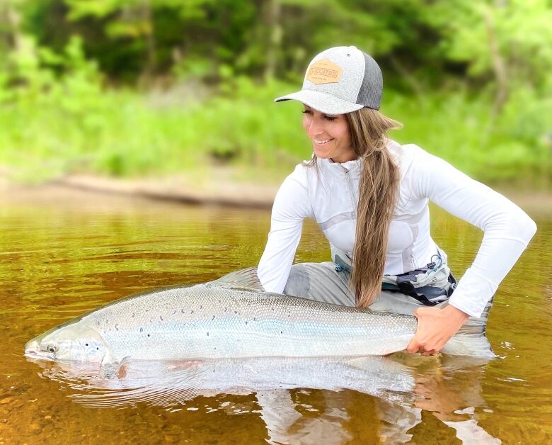 A woman in a white shirt and a baseball hat stands in a river holding a large salmon.