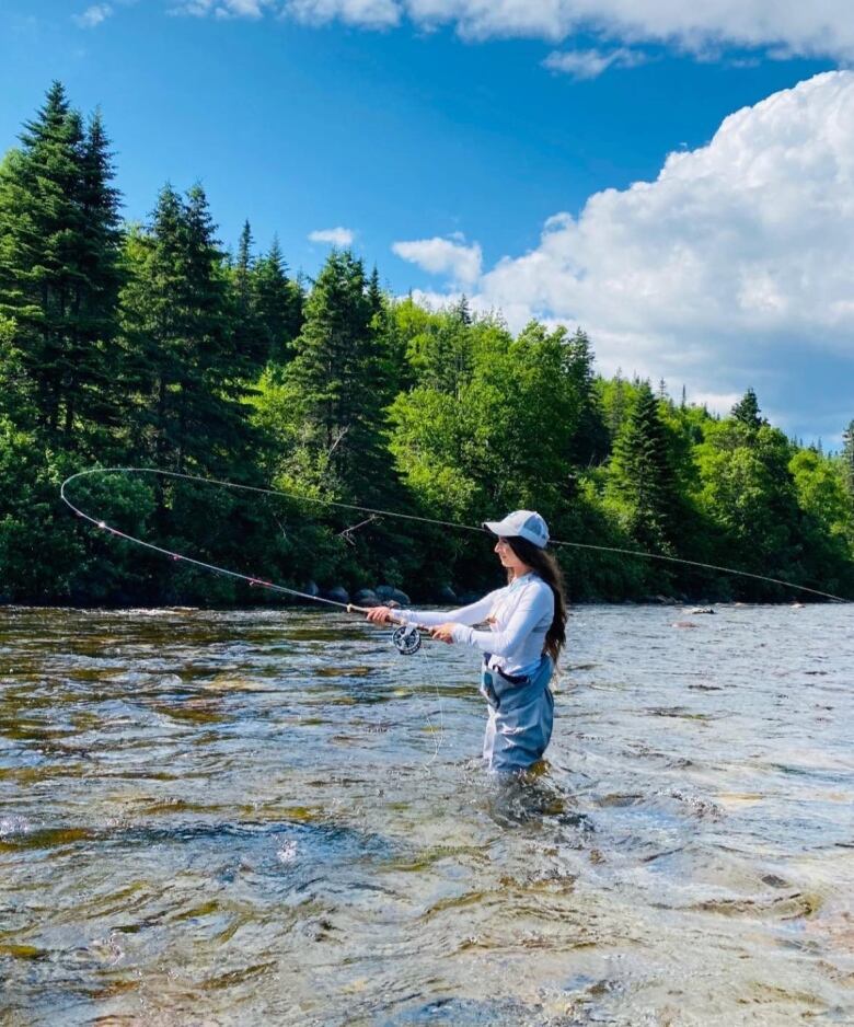 A woman stands in a river, trees behind her, casting a long line.