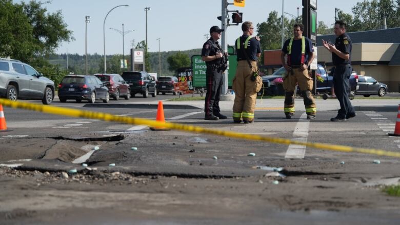 firefighters standing with police officers. there is a broken hole in the road and caution tape around it.