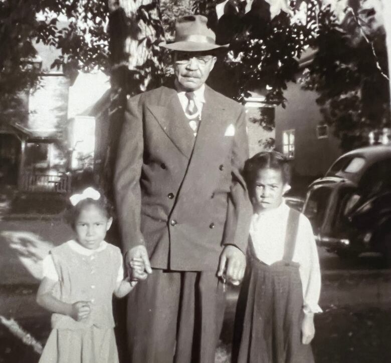 Black and white image of a man in a suit and brimmed hat standing with two young girls.