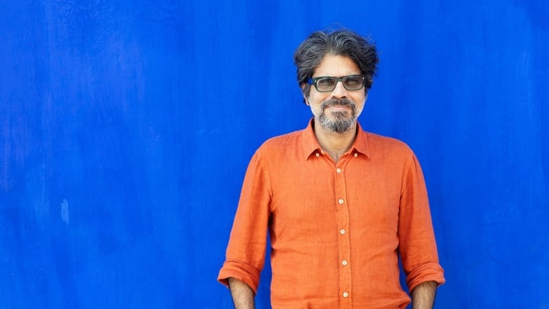 An Indian man with a greying hair, beard and glasses smiles in front of a blue background.