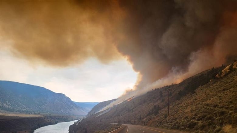 Flames and smoke arise from a hilltop next to a river.