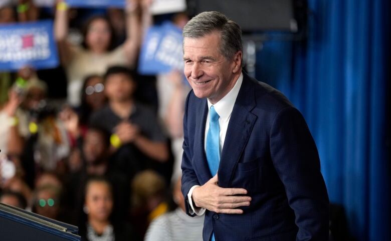 White politician on a podium in a suit and tie, taking a bow in front of a cheering crowd