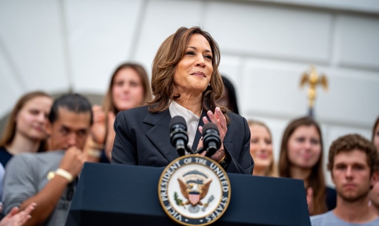 A woman in a navy suit with a grey shirt stands with her hands together at a podium marked with the seal for the vice president of the United States.