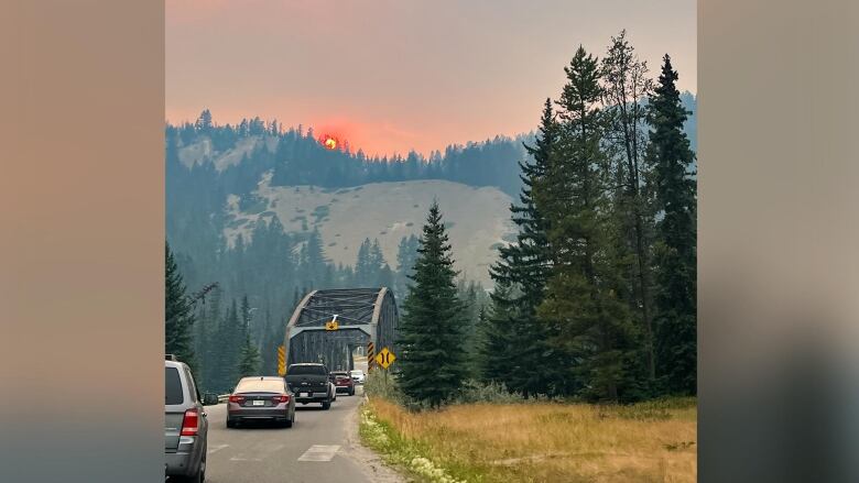 Cars on a single lane road and bridge under a hazy sky and red sun from wildfire smoke.