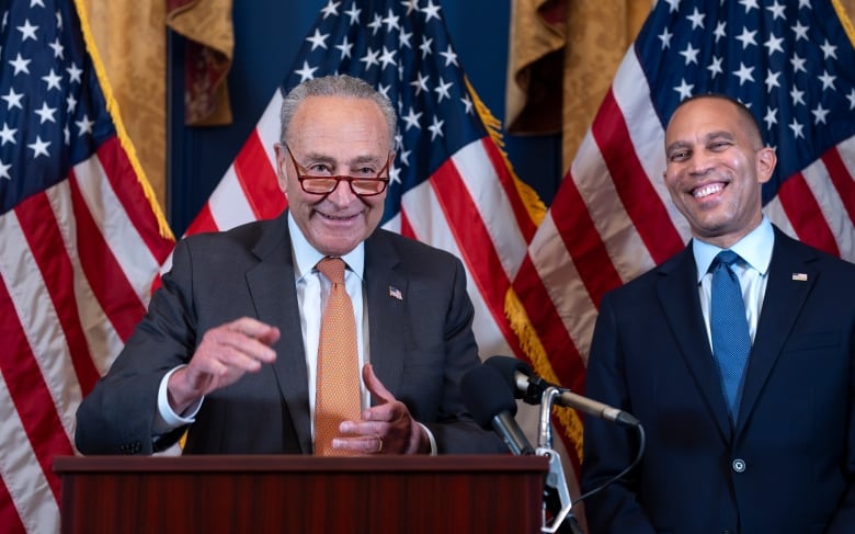 Two men in suits, one with a peach tie and one with a blue tie, stand in front of large American flags at a news conference.