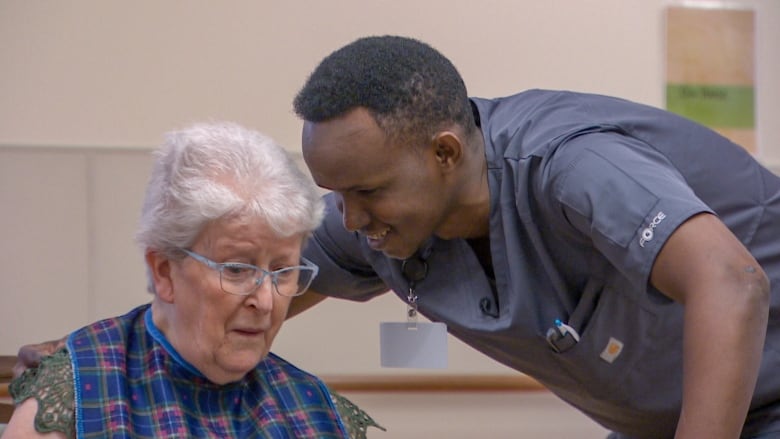 Man wearing scrubs leans over elderly woman wearing a bib. 