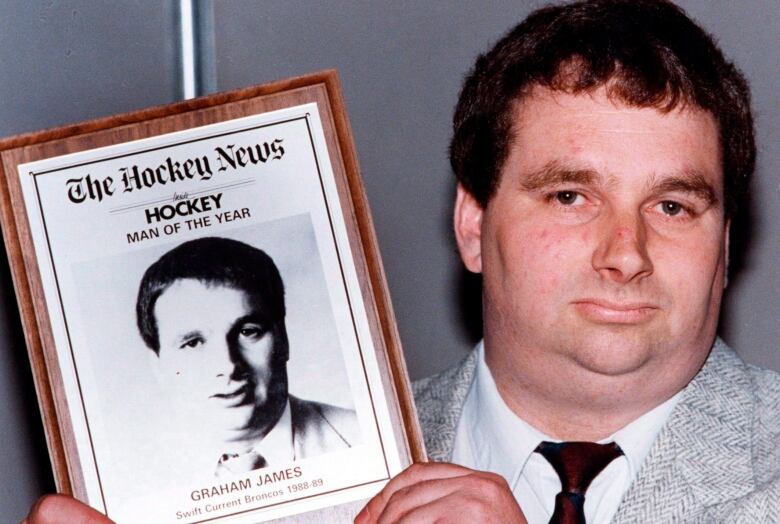 A man holds an award to the camera that says 'The Hockey News, Hockey, Man of the Year, Graham James.'