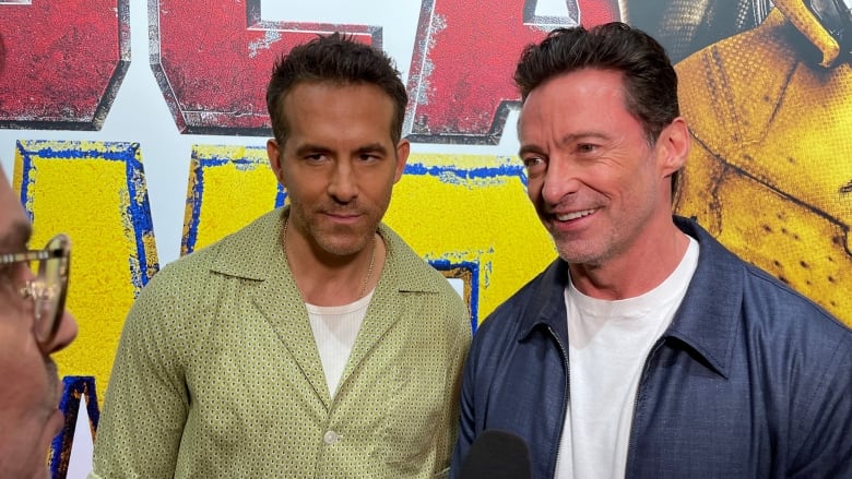 Ryan Reynolds, on the left wearing a green top, and Hugh Jackman, on the right right wearing a blue bomber, speak  on the red carpet.