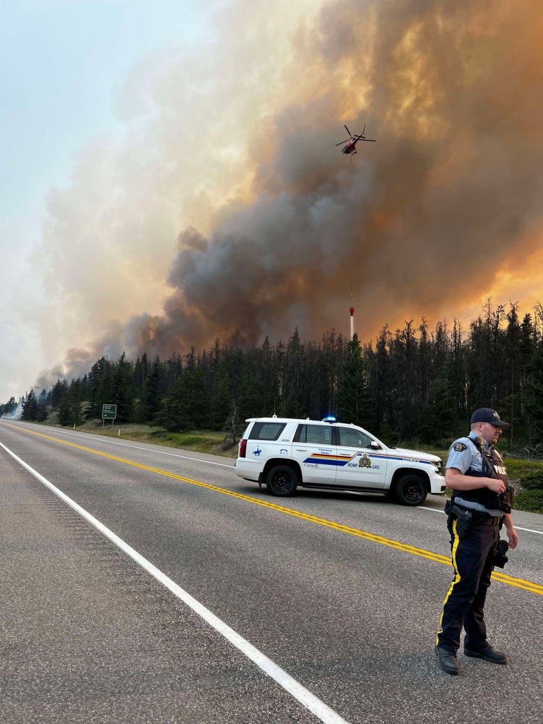 A man in a police uniform stands in front of an RCMP vehicle parked across a highway road. 