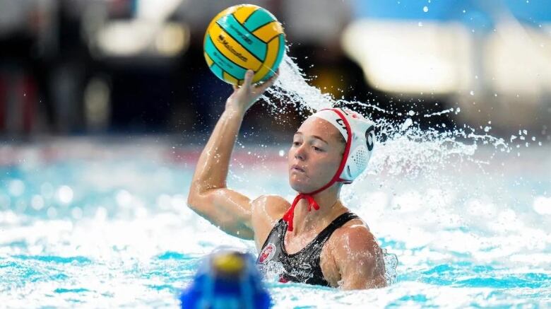 A woman in a pool holds up a water polo ball.