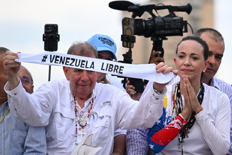 An older cleanshaven man holds a banner that says 'Venezuela Libre' as a woman with her hair pulled back looks on to his left.