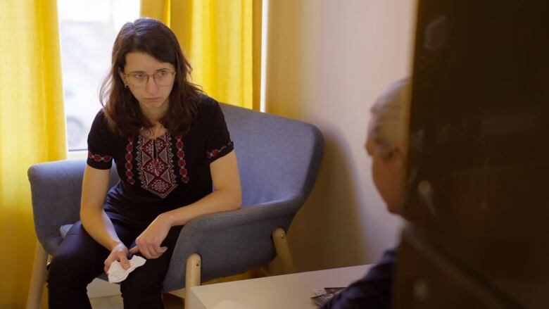 A young woman with dark hair sits on a chair leaning forward. She is listening to a blonde woman who is in the foreground with her back to the camera. 