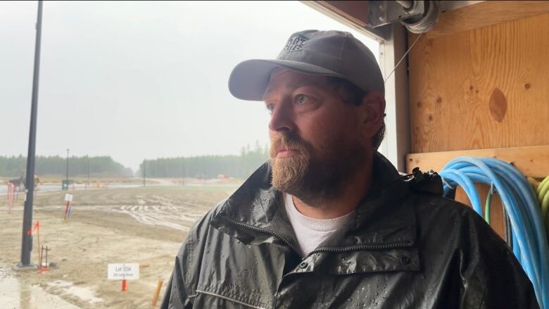 A man in a rain jacket and grey hat stand in front of a construction site.