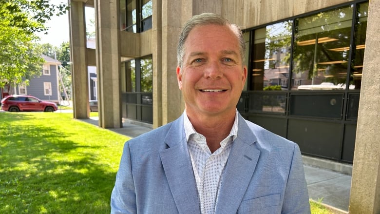 A man in a white shirt and grey blazer looks at the camera.