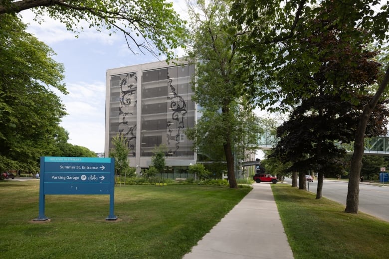 A grey, multi-storey parking garage is seen in the distance surrounded by trees, green grass and a sidewalk.