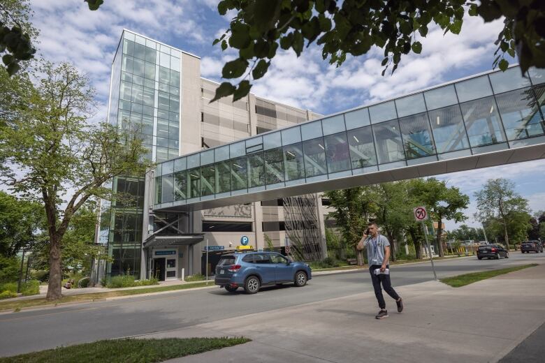 A person talks on a cellphone while walking down the sidewalk in front of a grey, multi-storey parking garage with a pedway. A blue car is on the street in front of the parkade.