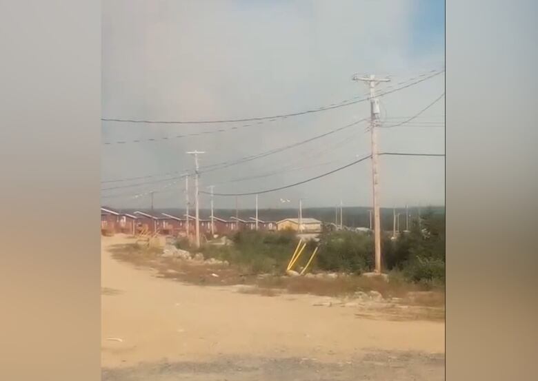 An unpaved road stands in front of a group of red houses. Smoke covers the sky.