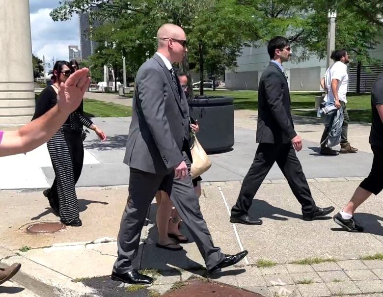 A man in a suit wearing sunglasses walks on a sidewalk with supporters.