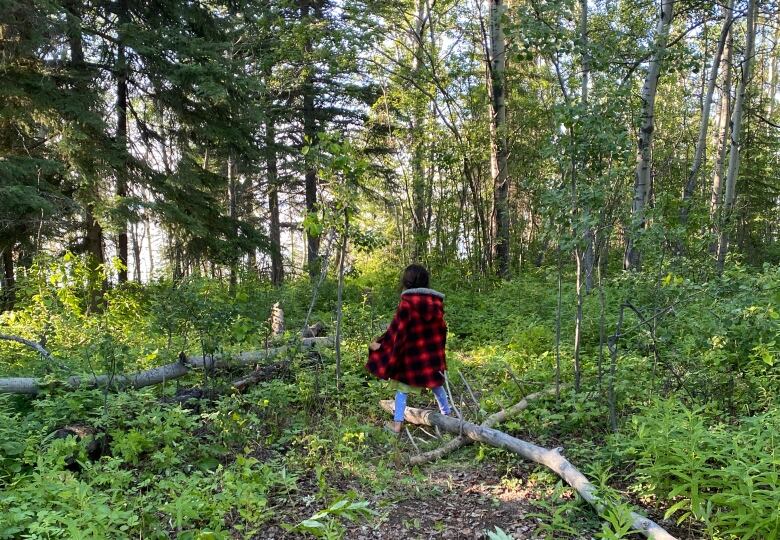 A girl walks through a forest, wearing a poncho and rainboots.