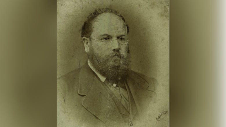 A sepia portrait of a portly and well-dressed man wearing wire-rimmed glasses.