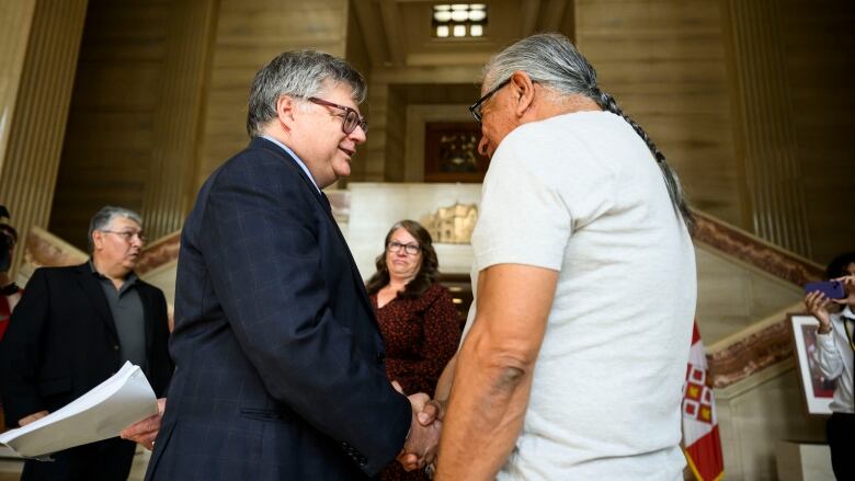 A First Nation chief with long grey hair tied back in a braid shakes hands with a man with short grey hair wearing a blue suit inside the foyer of the Supreme Court of Canada. 