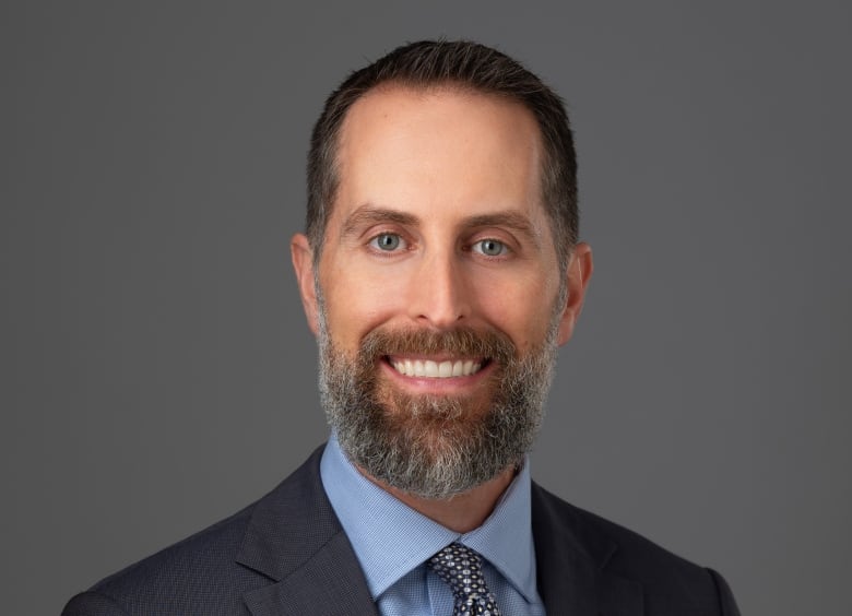 A man with a suit and tie and a beard smiles at the camera.